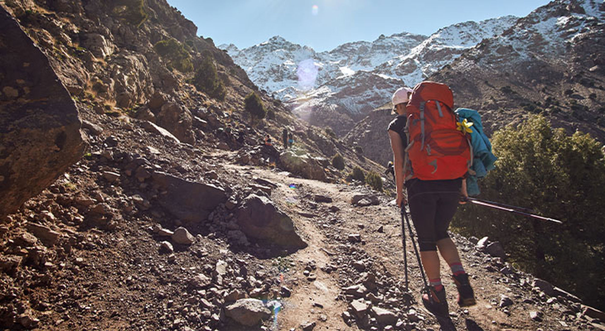 Mount Toubkal Wandelaar met trekkingstokken en rugzak op bergpad richting besneeuwde bergtoppen.