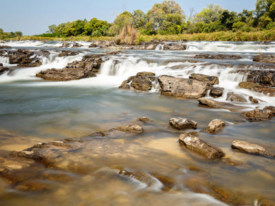 Stromend water over rotsen in een brede rivier met groene oever, op de route Kaapstad–Victoria Falls.