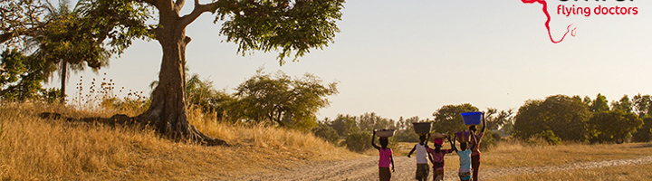 Kinderen lopen over een zandpad met bakken op hun hoofd, met het logo van Amref Flying Doctors.