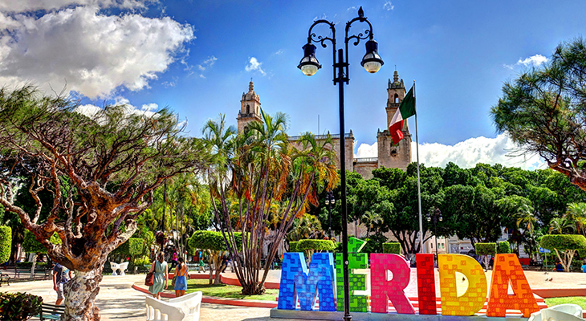 Mérida Kleurrijk Mérida-bord in een groen stadspark met kathedraal en Mexicaanse vlag op de achtergrond.