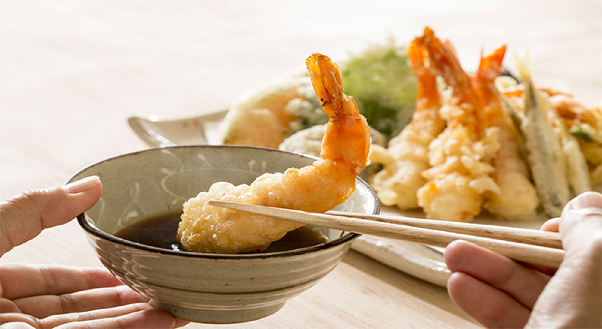 Gefrituurde garnalen Tempura garnaal wordt met eetstokjes in sojasaus gedoopt, met een bord tempura op de achtergrond.
