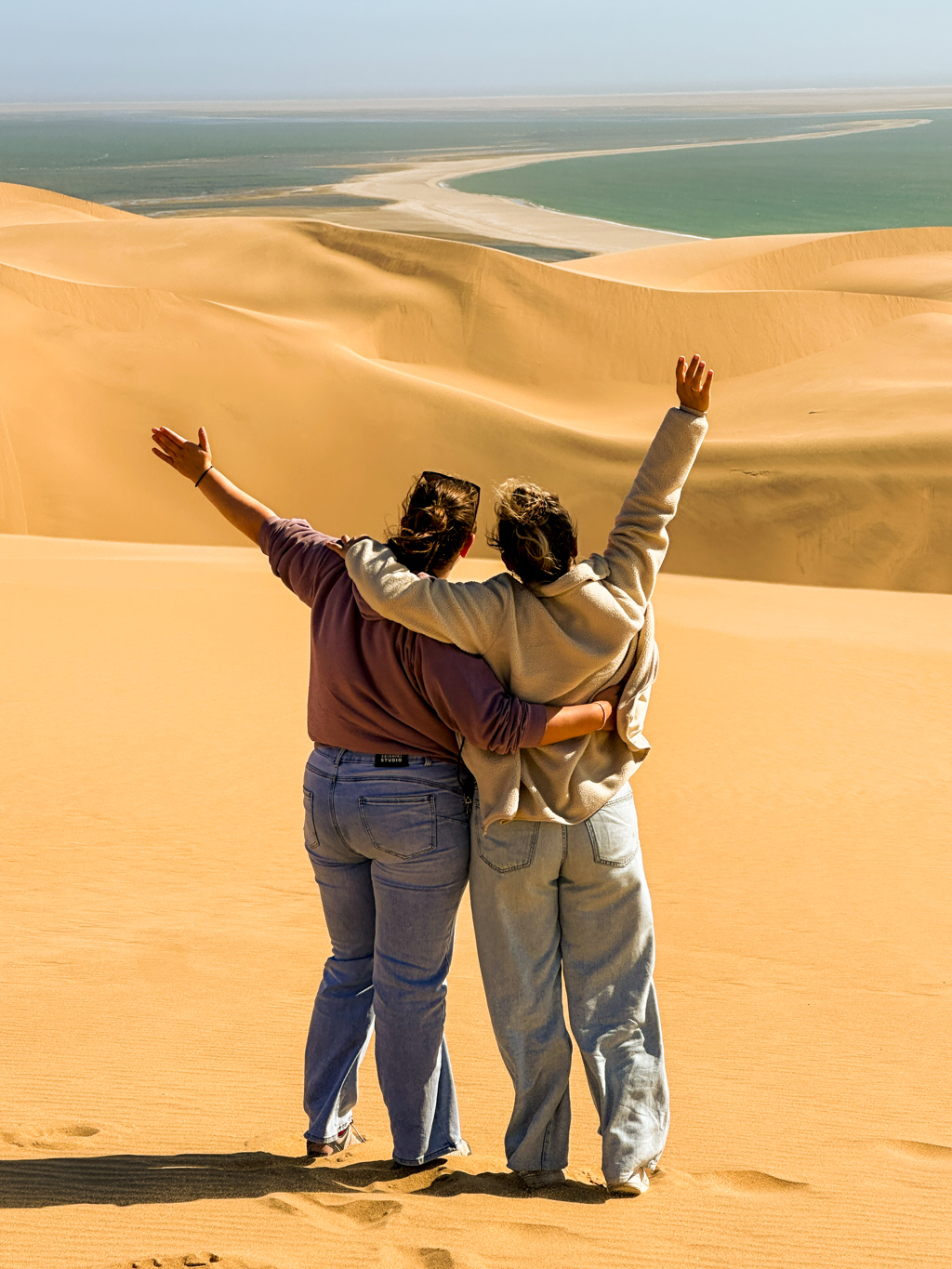 Twee reizigers met opgeheven armen op zandduinen met uitzicht op de kust tijdens 22-35ers reis Zuidelijk Afrika.