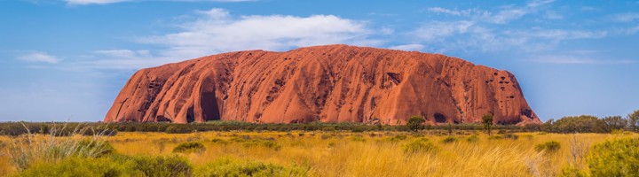 Uluru rotsformatie in de Australische Outback, omringd door rood zand en gras onder een blauwe lucht.