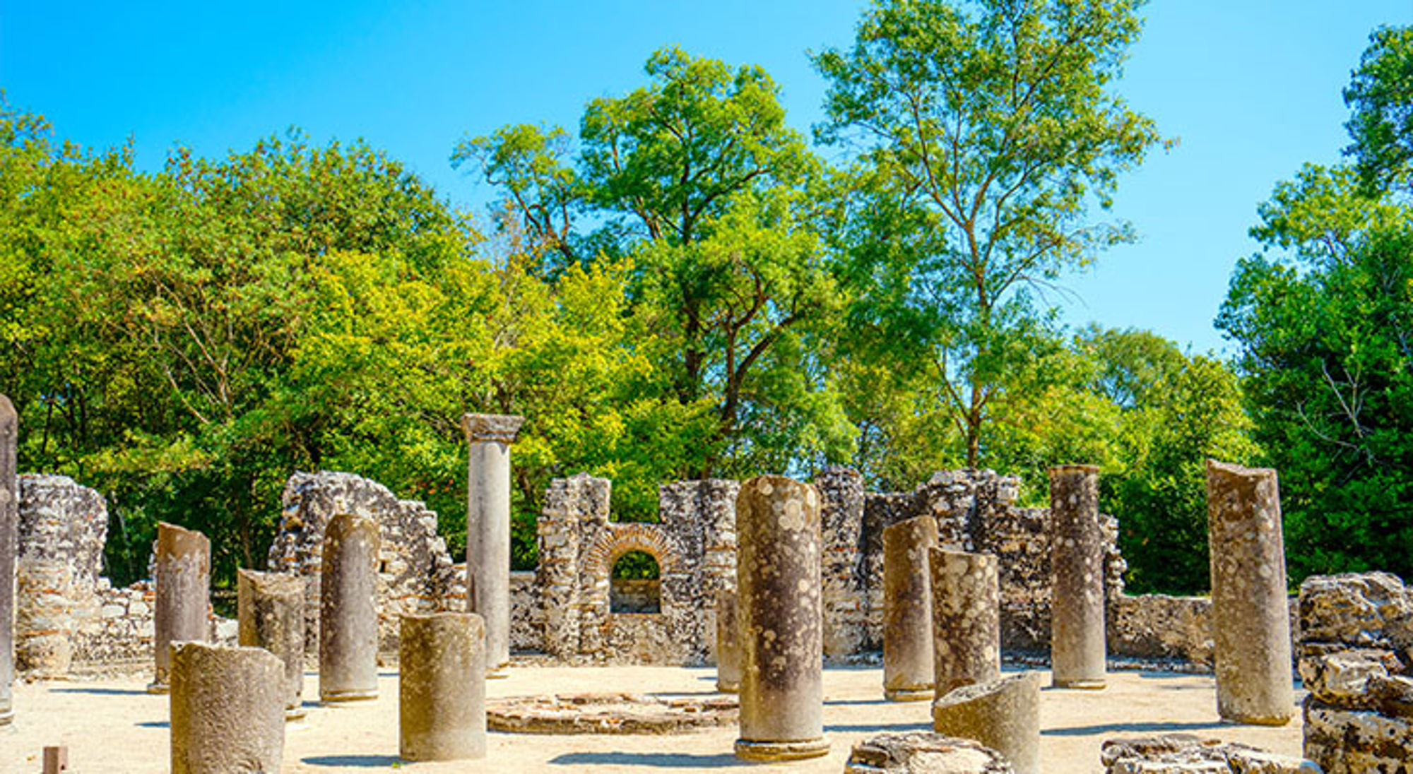 Butrint Ruïnes met stenen zuilen tussen groene bomen onder een heldere blauwe lucht.