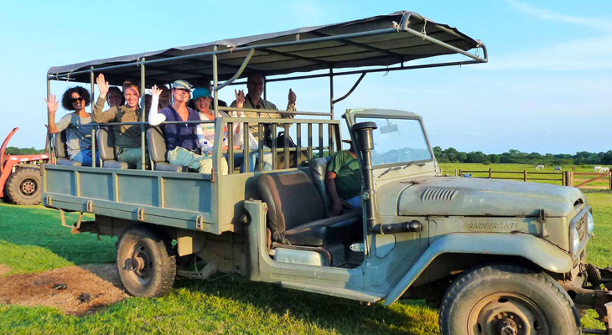 Groep reizigers zwaait vanuit een open safaritruck op een groene vlakte.