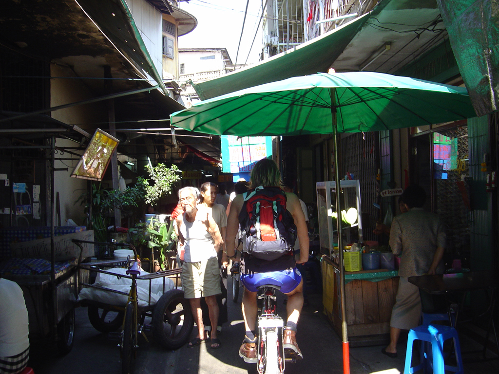 Fietser met rugzak door smalle straat in Thailand, langs marktkraampjes en groene parasol.