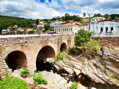 Stenen boogbrug over een rotsachtige rivier met kleurrijke huizen in een Braziliaans stadje op de Route Brazilië.