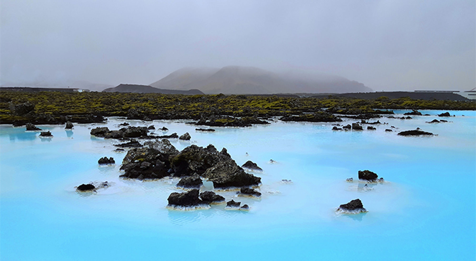 IJsland Stoomend blauw geothermisch meer met lavastenen en mos, met mistige bergen op de achtergrond.