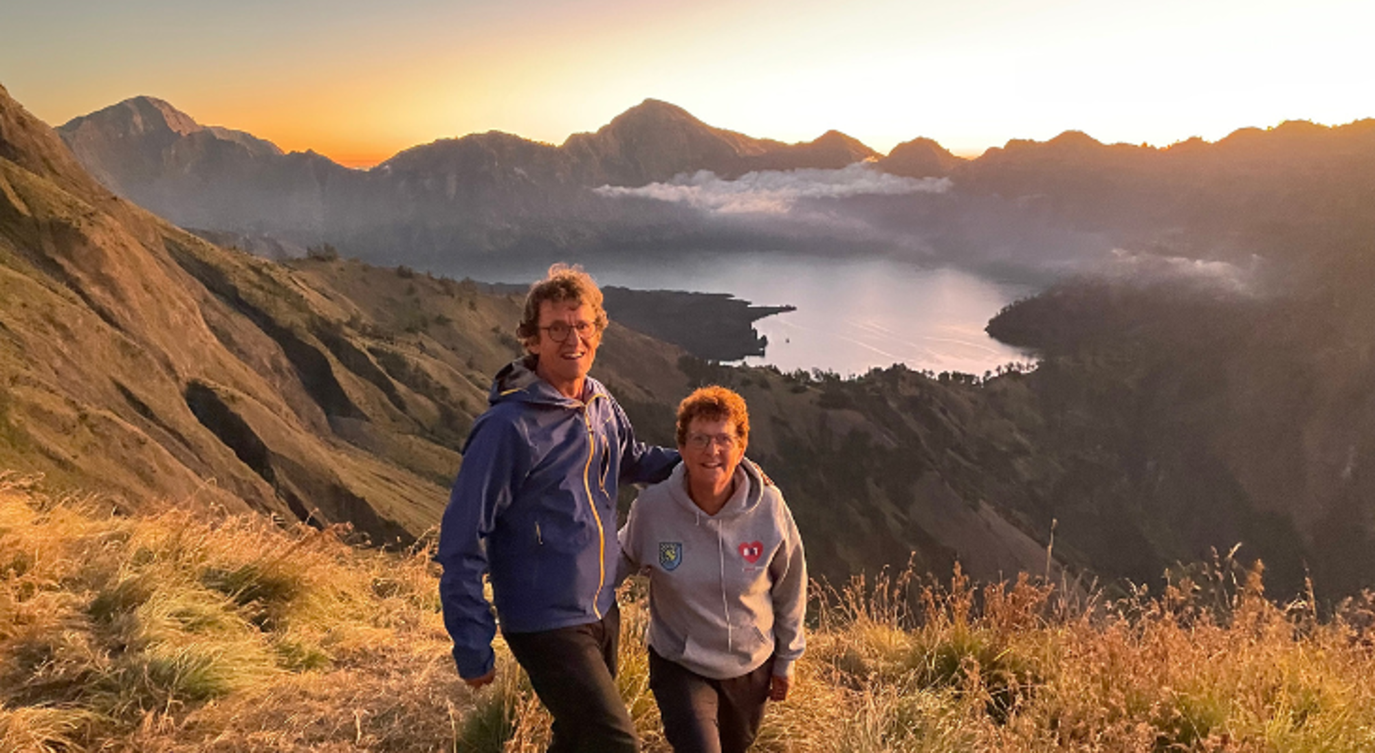 Mount Rinjani op Lombok Twee wandelaars op een berghelling met uitzicht op een kratermeer en bergen bij zonsondergang.