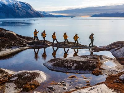 Wandelaars in gele jassen langs de kust van Groenland, met fjord, ijsbergen en bergen op de achtergrond.