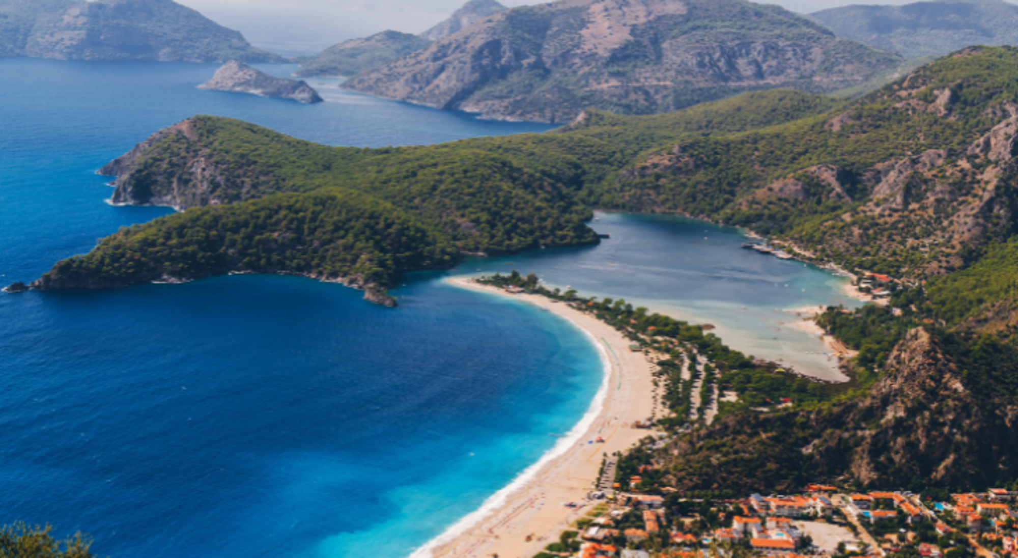 Panoramisch uitzicht op een halvemaanvormig zandstrand met turquoise zee, lagune en groene bergen.