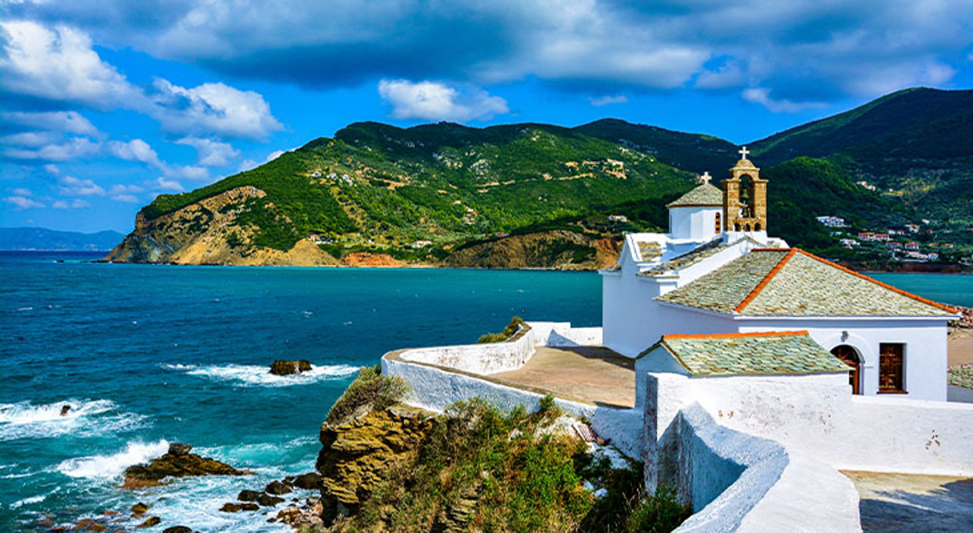 Skopelos Witte kapel aan de kust met uitzicht op zee en groene bergen onder een blauwe lucht.