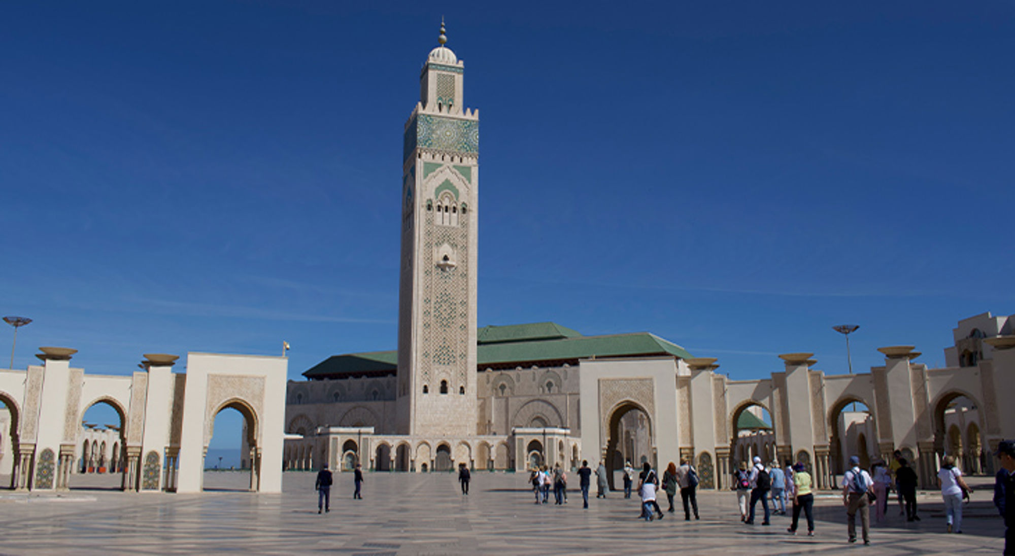 Hassan II Moskee Hassan II-moskee in Casablanca met hoge minaret en bezoekers op het plein onder een heldere blauwe lucht.