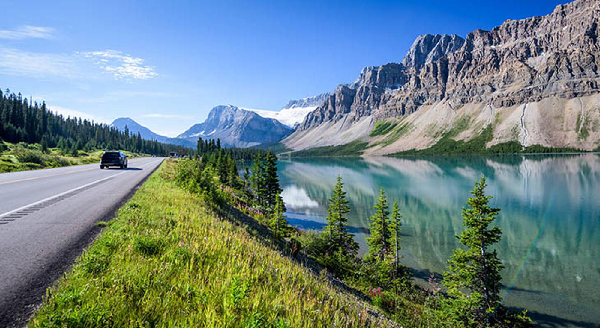 Icefields Parkway in Canada Auto op bergweg langs turquoise meer met spiegelende bergen en naaldbomen onder heldere blauwe lucht.