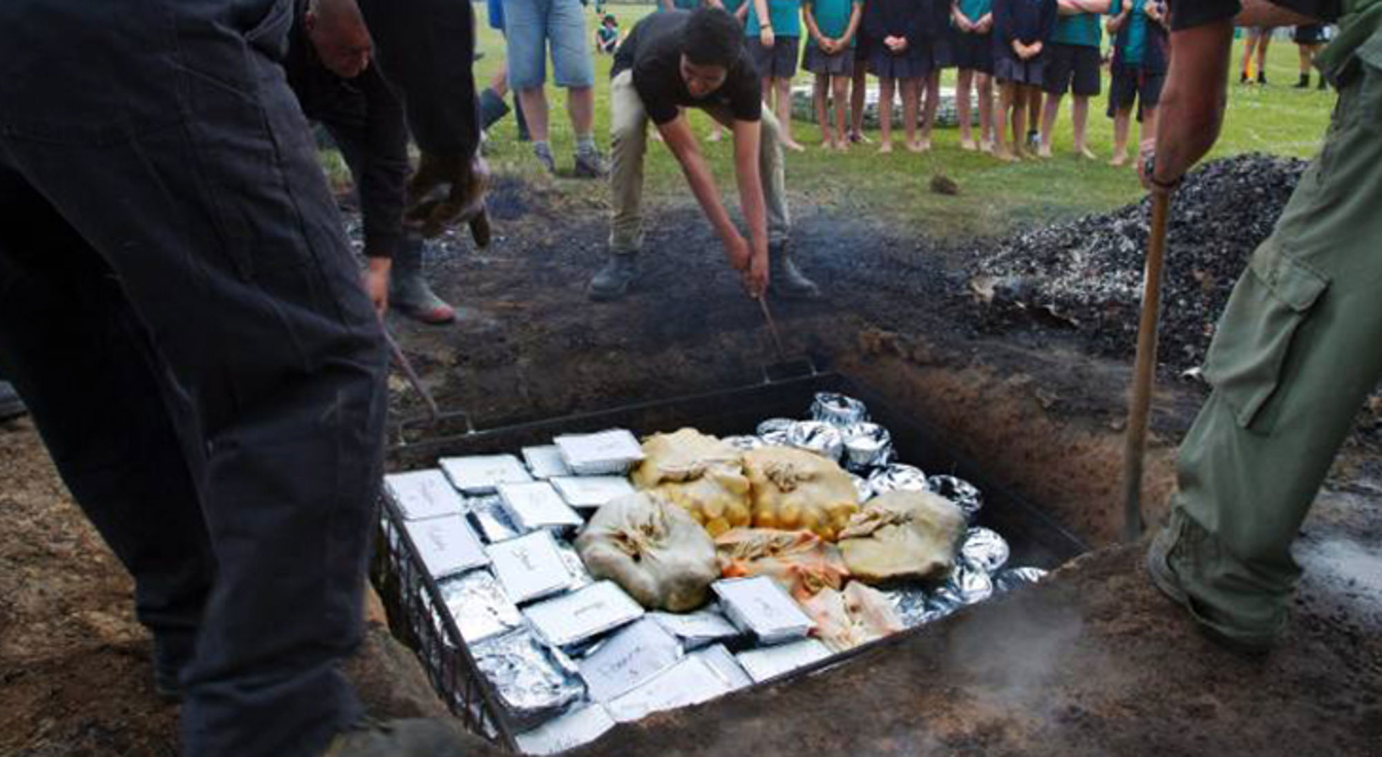 Koken in een kuiloven met pakketten in folie, terwijl deelnemers het vuur verzorgen.
