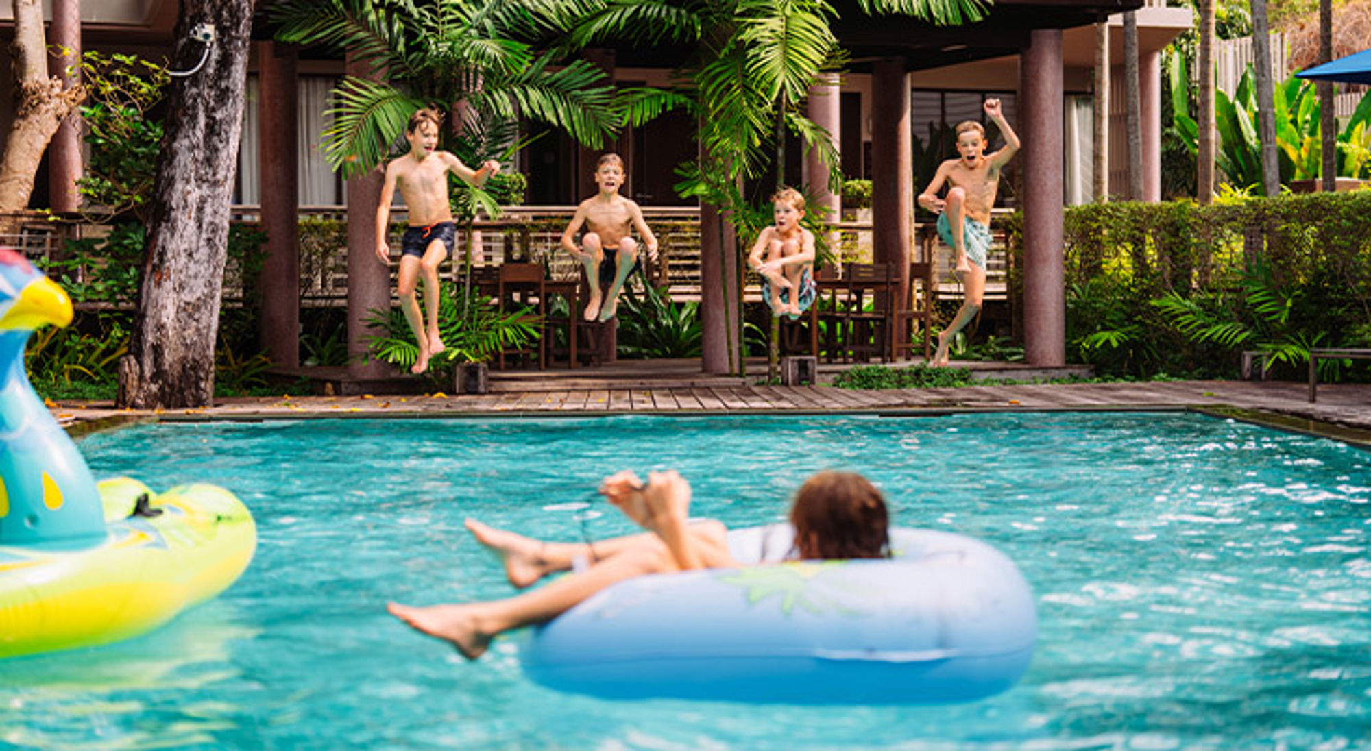 Kinderen springen in een zwembad bij een resort, terwijl iemand ontspant op een zwemband.