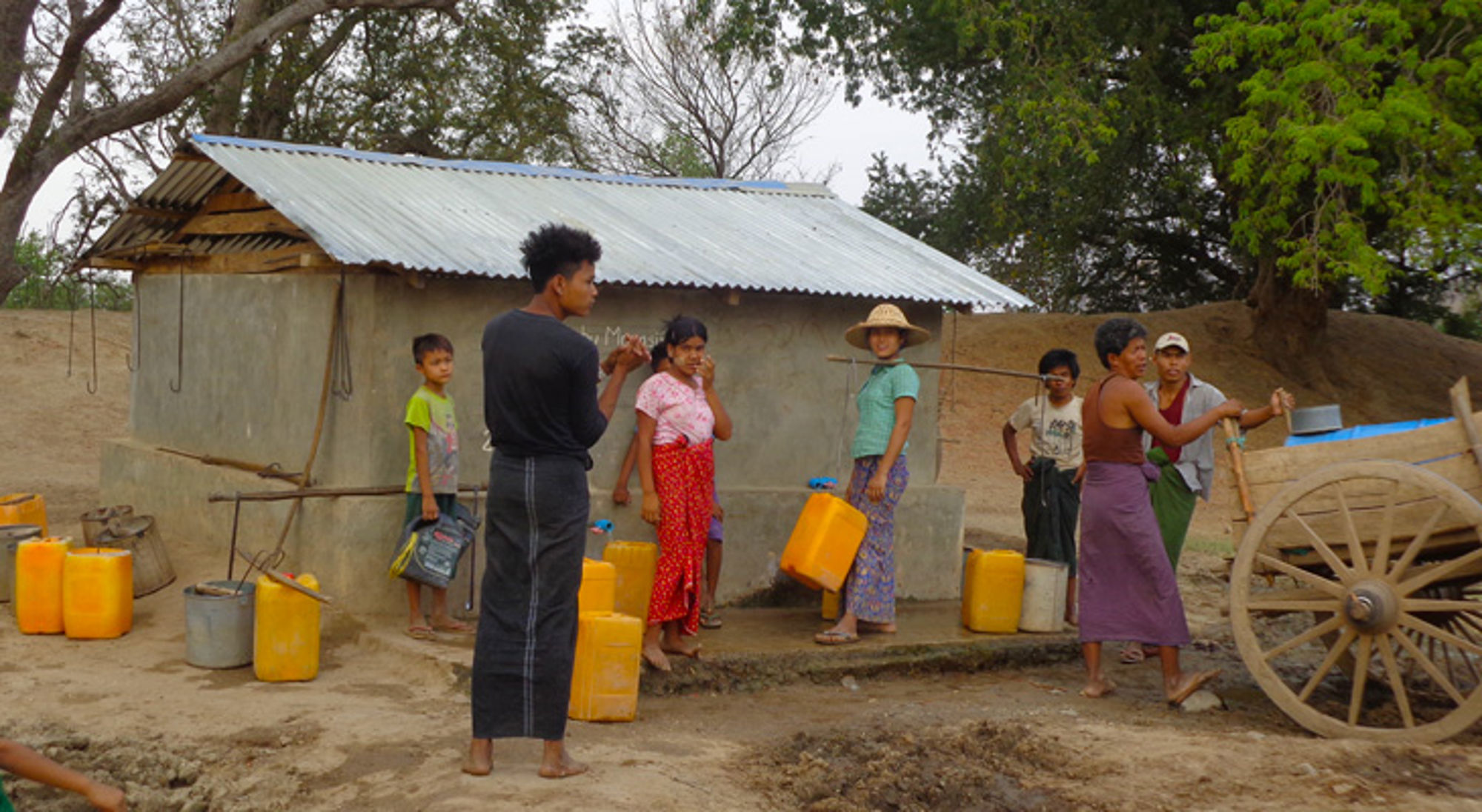 Lokale bevolking in Myanmar Lokale bewoners vullen gele jerrycans bij een waterpunt naast een klein gebouw en houten kar.