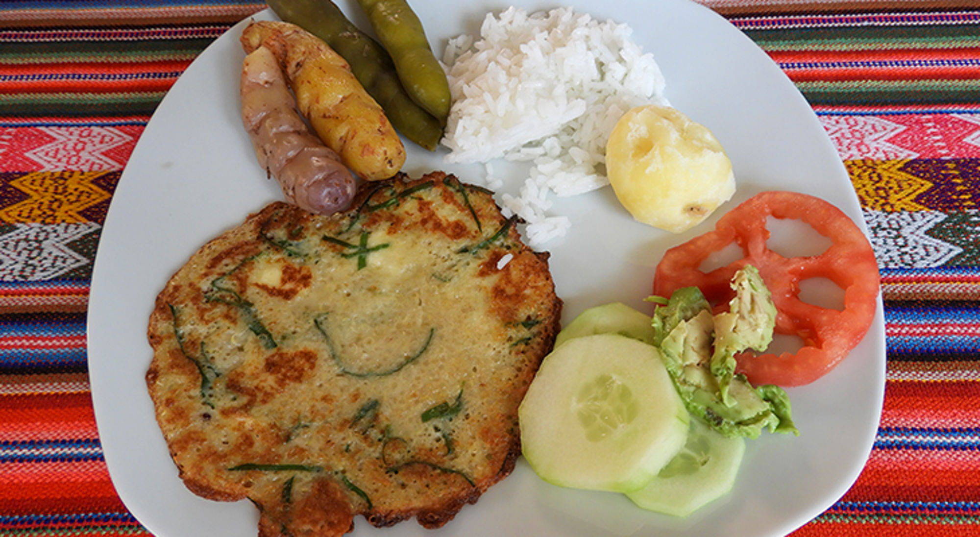 Bord met hartige pannenkoek, rijst, worst, aardappel, bonen en salade op kleurrijk tafelkleed.