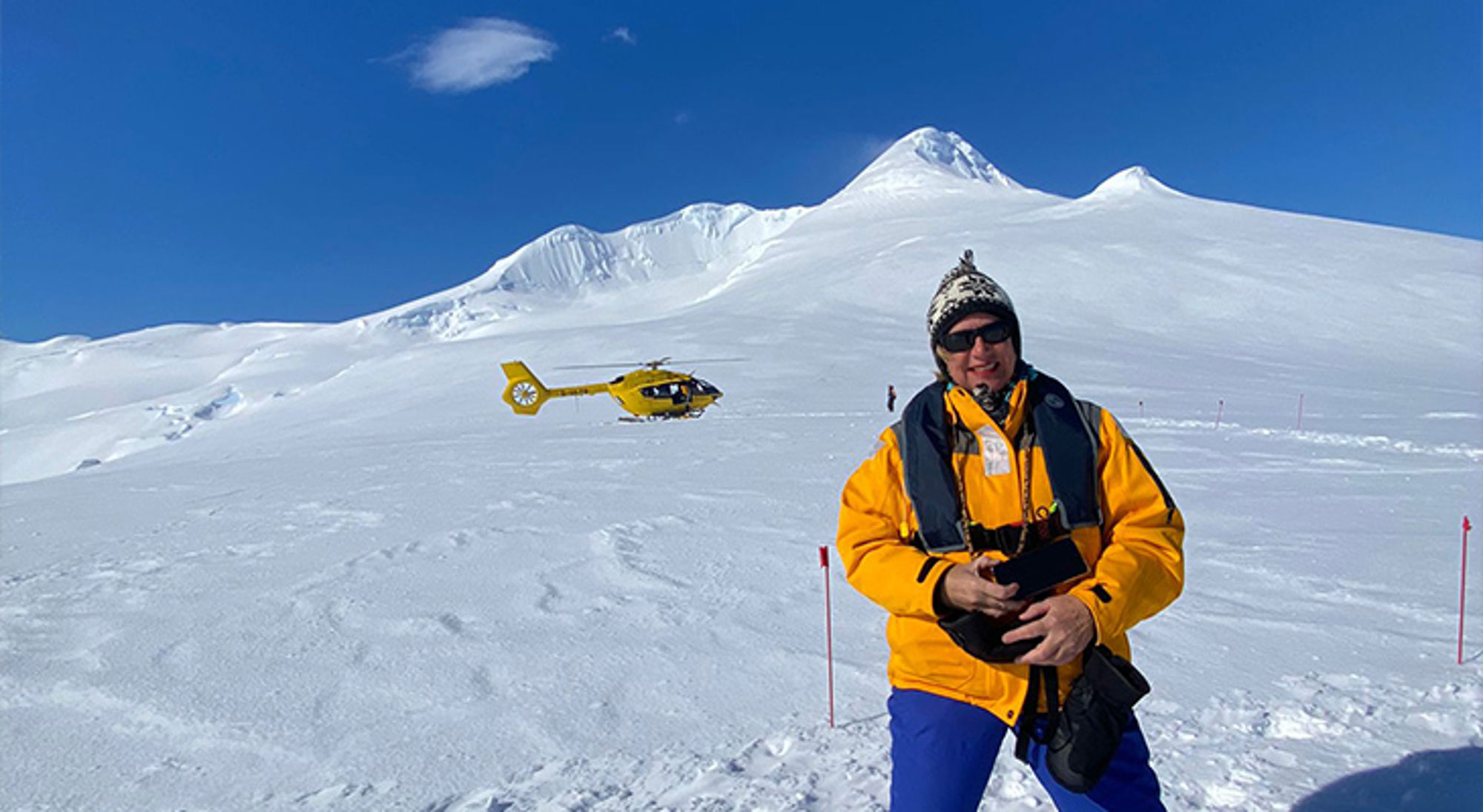 Helilanding Antarctica Reiziger in winterkleding op een besneeuwde berg, met een gele helikopter op de achtergrond.