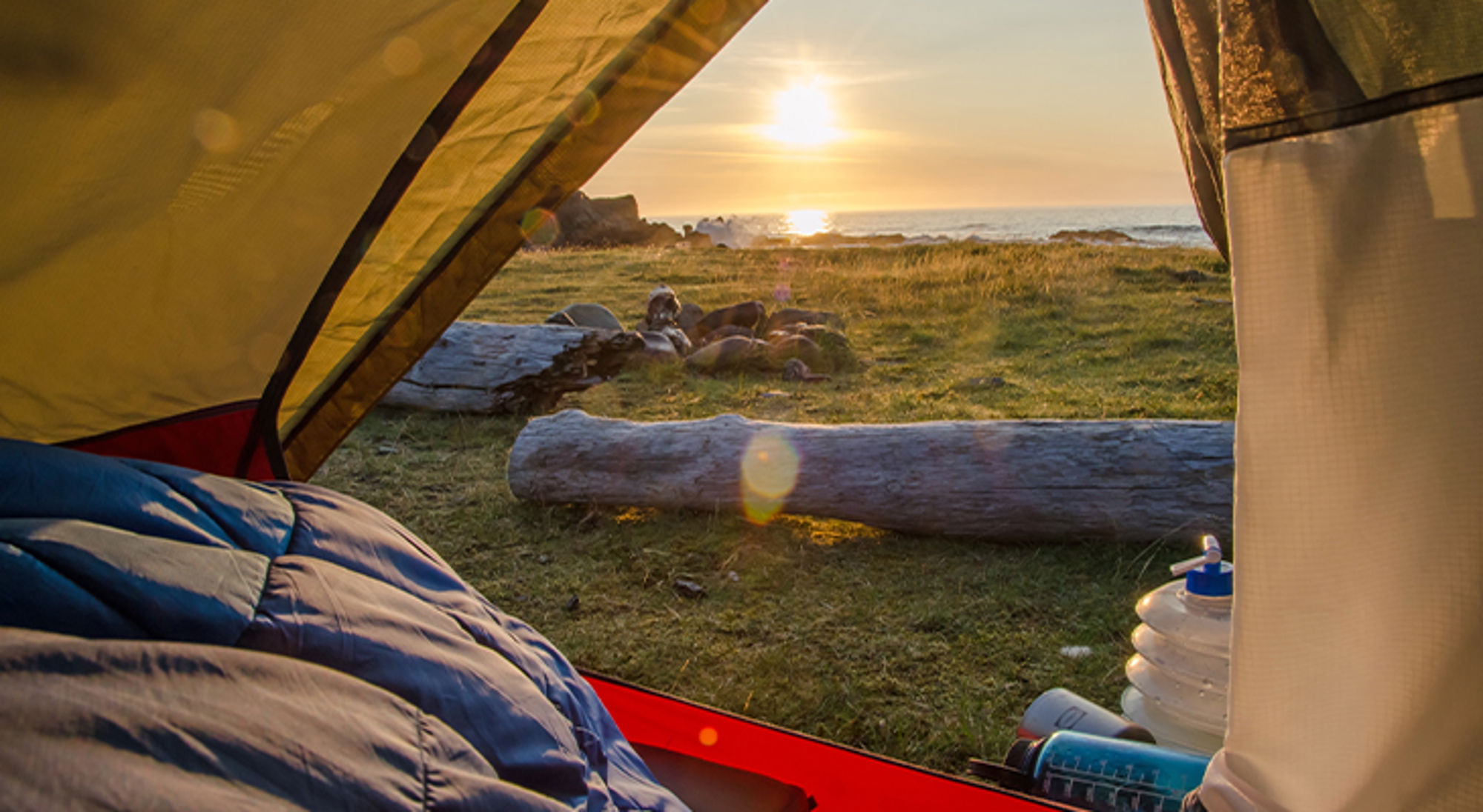 Kamperen IJsland Uitzicht vanuit een tent op een kustcamping bij zonsondergang, met grasveld, houtblokken en zee op de achtergrond.