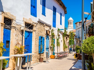 Straat in Cyprus met witstenen huizen, blauwe luiken en terrasjes, met kerktoren op de achtergrond.