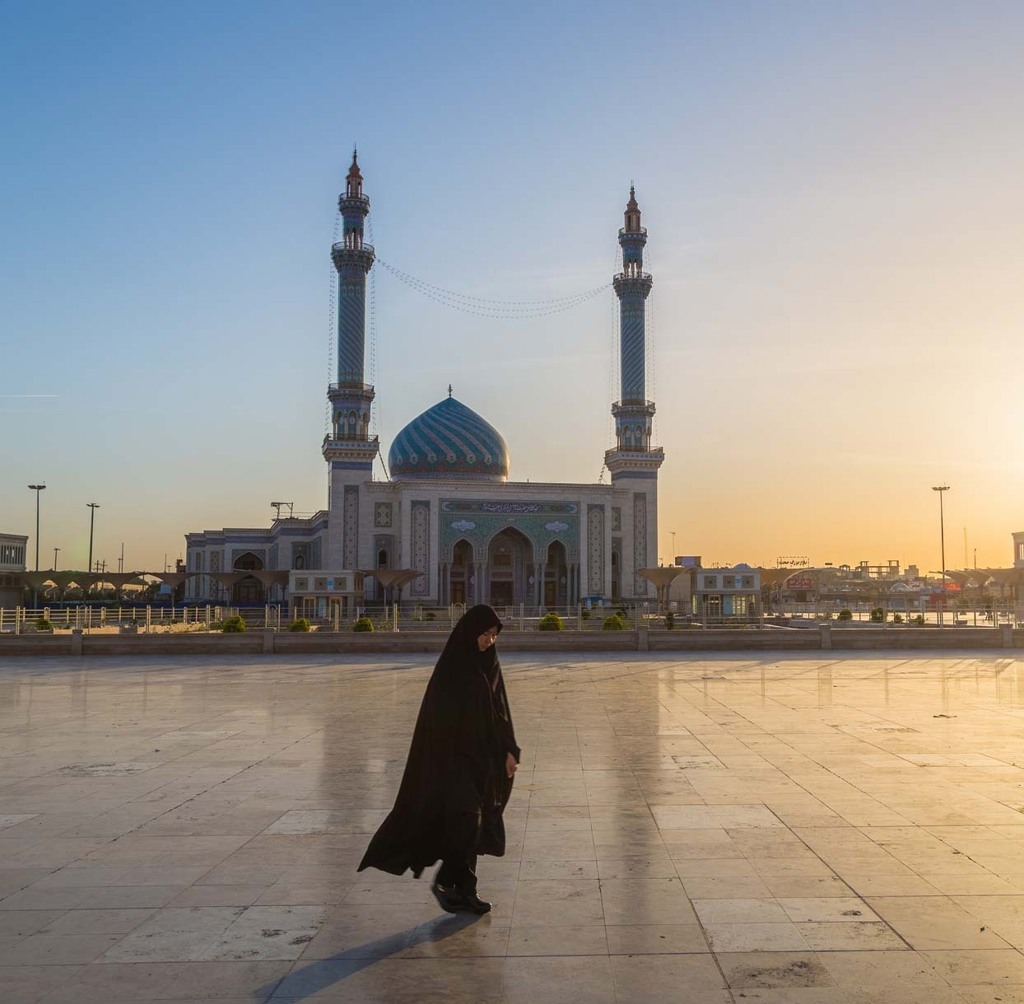 Vrouw loopt over plein bij moskee met blauwe koepel en minaretten in Iran, tijdens zonsondergang.