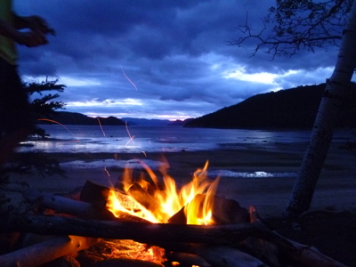 Kampvuur aan de oever van een meer in Canada bij schemering, met bergen en donkere wolken op de achtergrond.