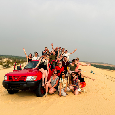 Groep 22-35ers poseert bij rode jeep in de zandduinen tijdens groepsrondreis Vietnam.