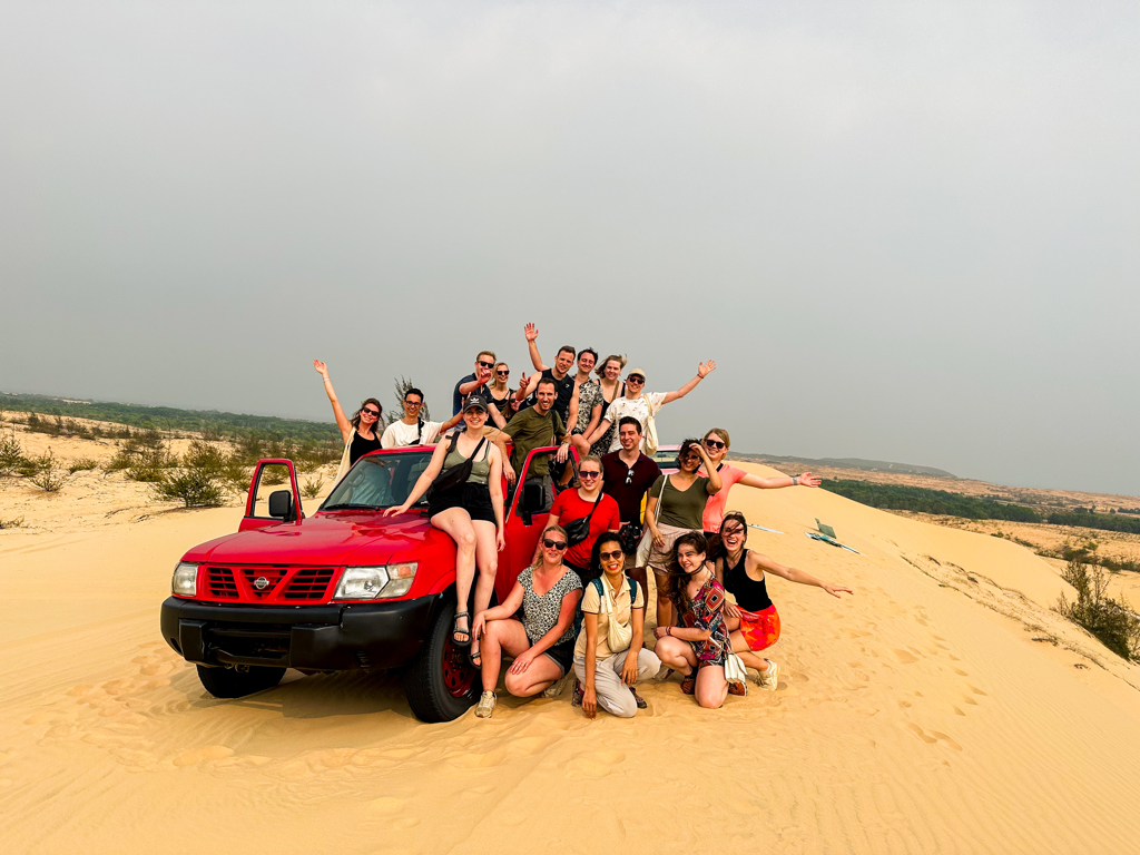 Groep 22-35ers poseert bij rode jeep in de zandduinen tijdens groepsrondreis Vietnam.