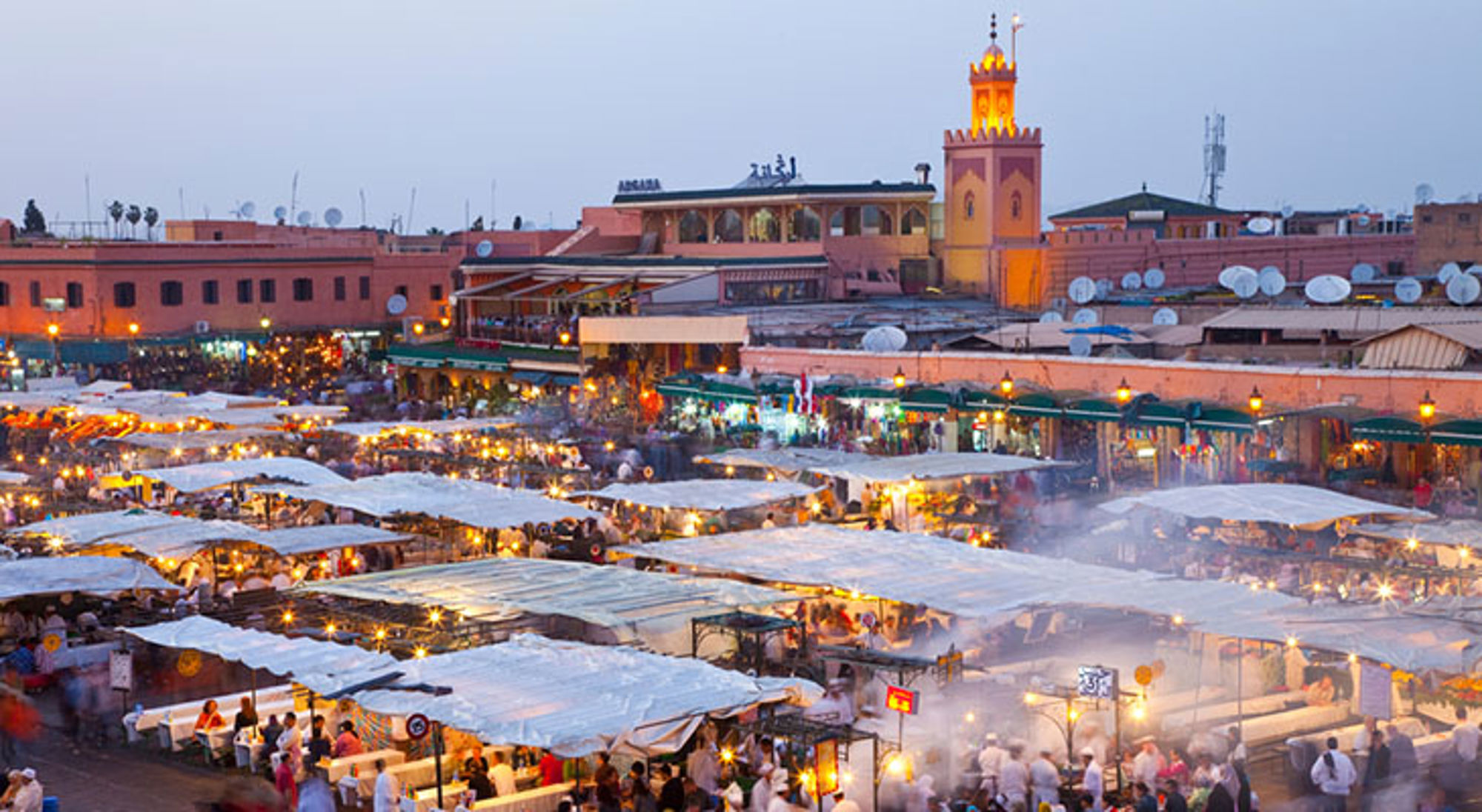 Djemaa El Fna Jemaa el-Fna-plein in Marrakech bij schemering, met drukke markt en verlichte minaret op de achtergrond.