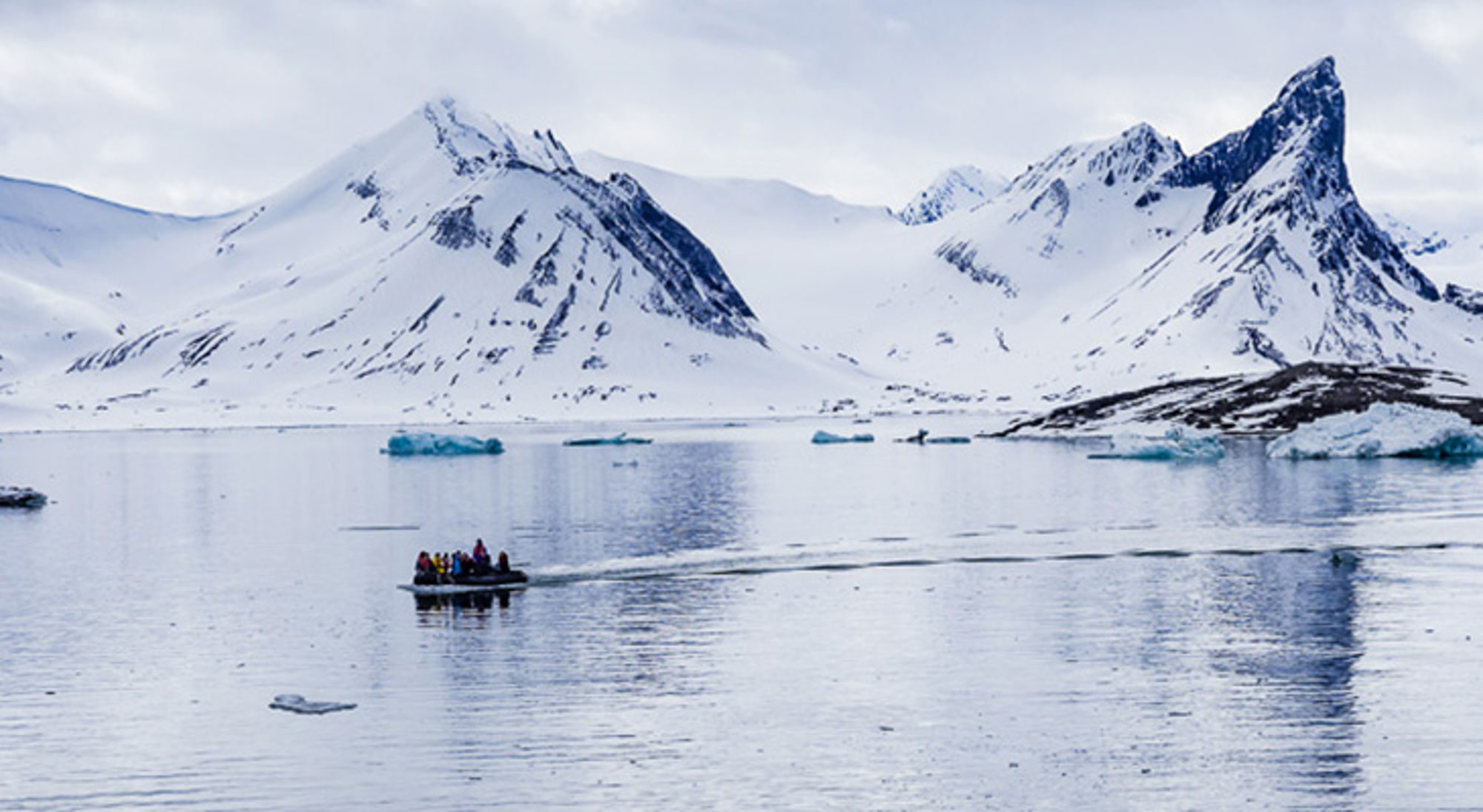 Zodiacboot met passagiers vaart tussen ijsbergen in een fjord met besneeuwde bergen op de achtergrond.