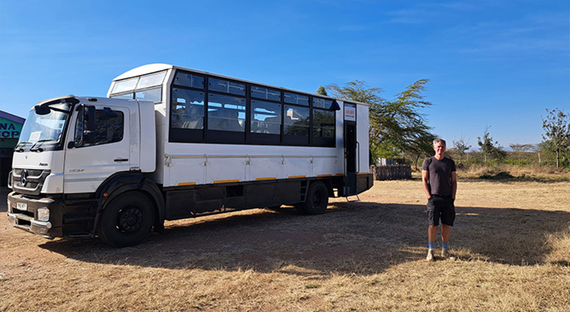 Safari truck met grote ramen geparkeerd op droge vlakte, met reiziger ernaast onder blauwe lucht.