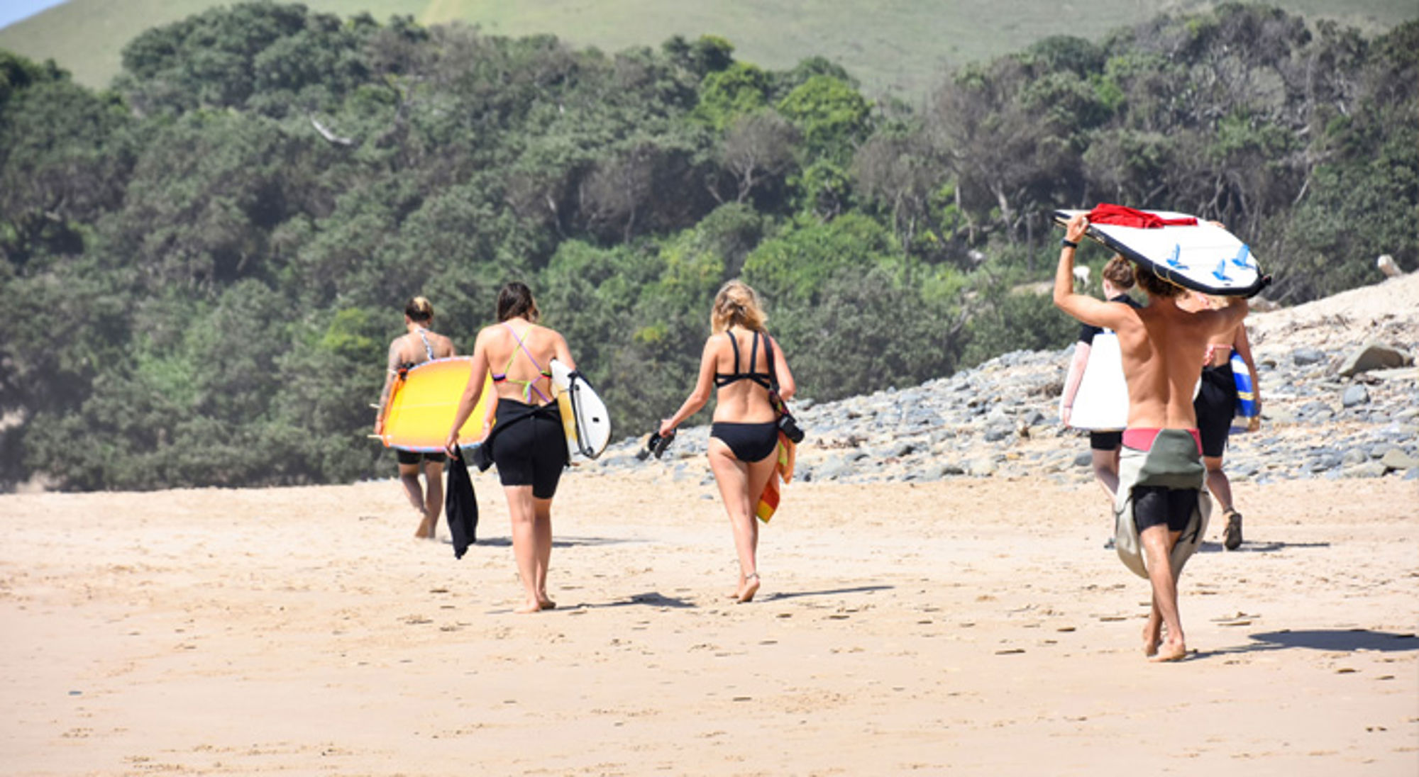 Surfers lopen met surfboards over een zandstrand in Zuid-Afrika, met duinen en groen op de achtergrond.