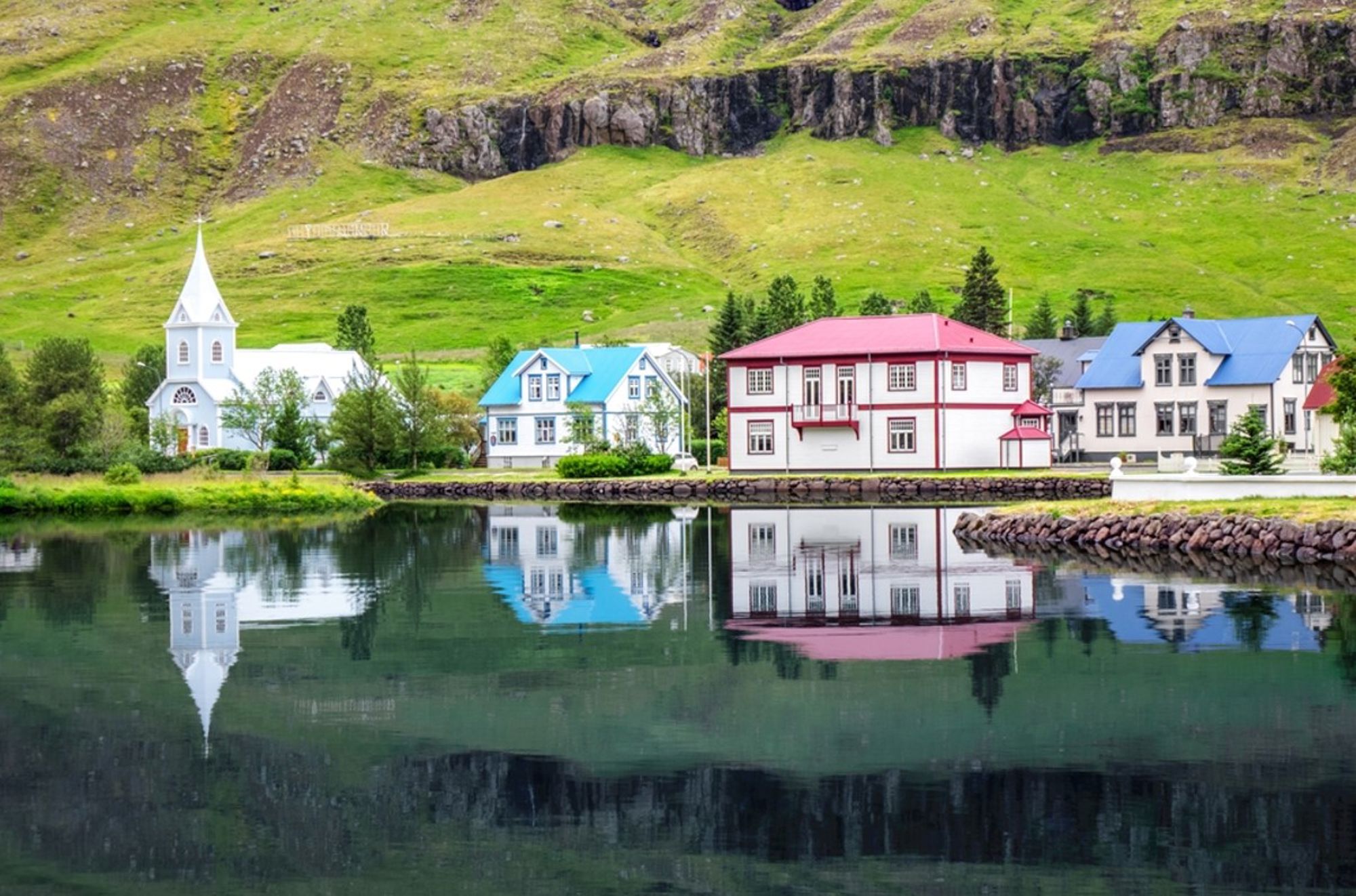 Kleurrijke huizen en witte kerk aan het water in Seyðisfjörður, IJsland, met weerspiegeling in de fjord.