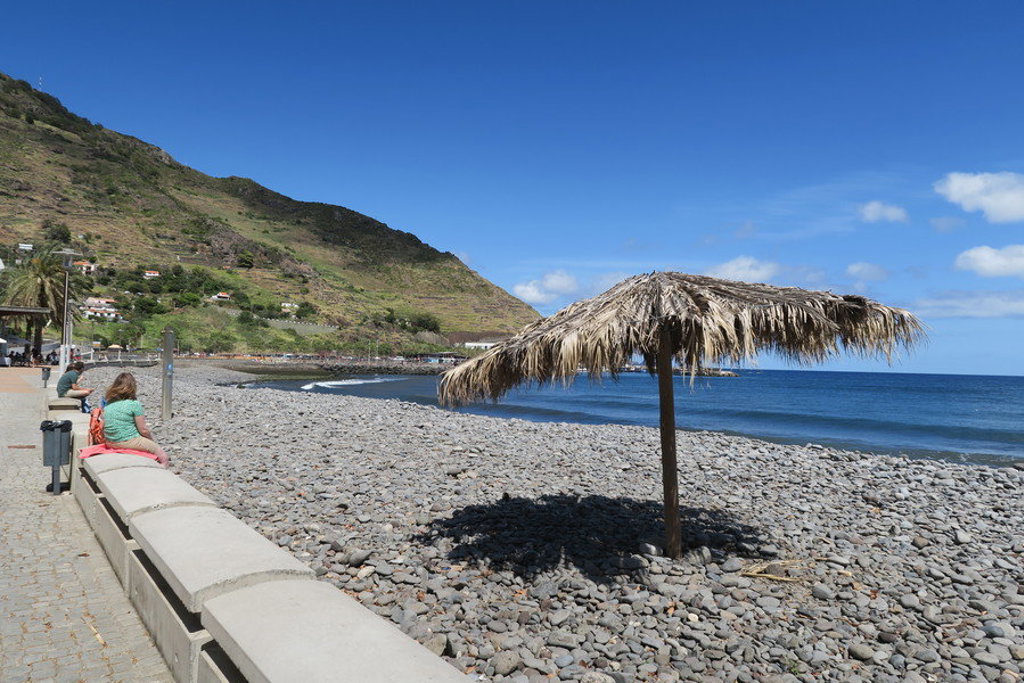 Kiezelstrand op Madeira met rieten parasol, promenade en uitzicht op de Atlantische Oceaan.