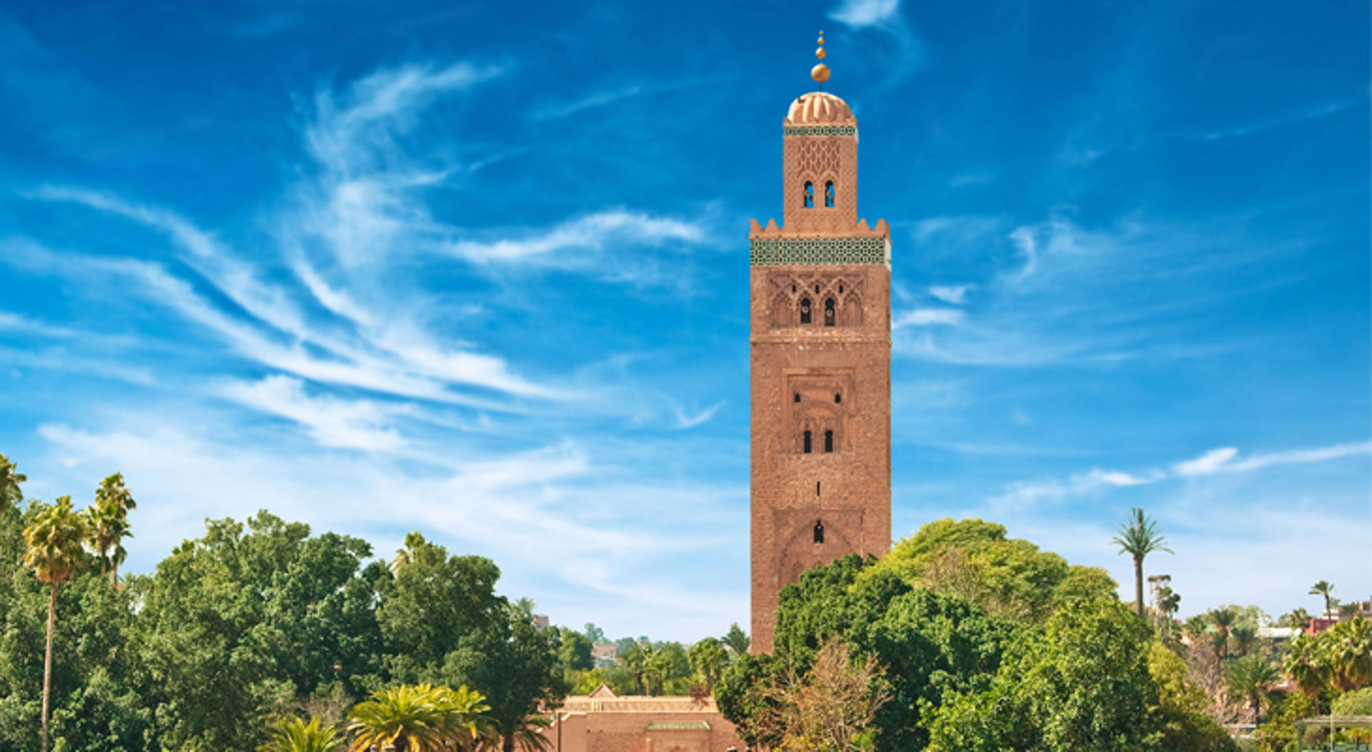 Minaret van de Koutoubia-moskee in Marrakech boven groene tuinen en palmbomen, onder een blauwe lucht.
