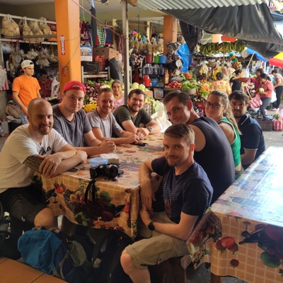 22-35ers groepsreis: reizigers aan tafel op een kleurrijke markt met groente- en fruitkramen in Guatemala.