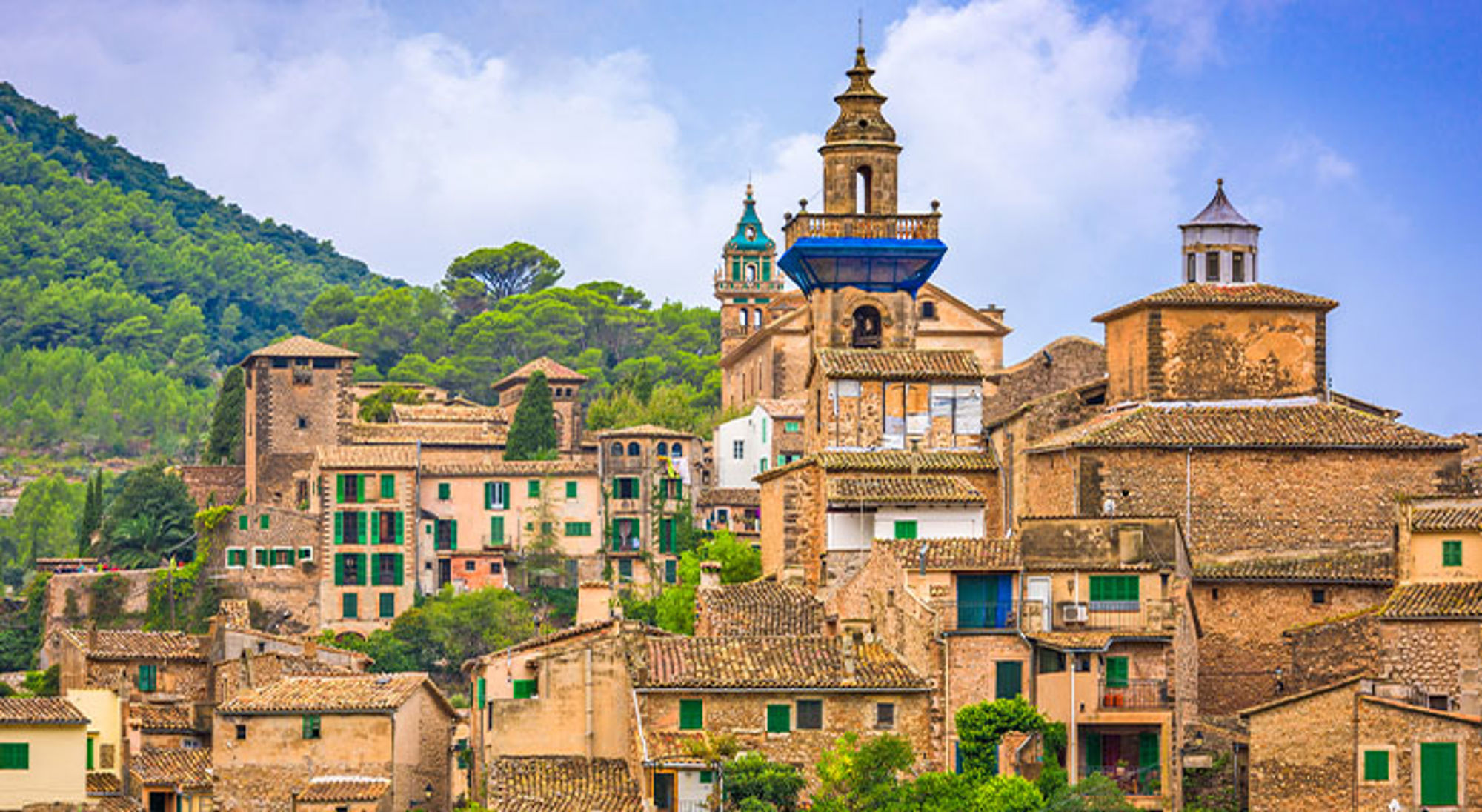 Mallorca Historisch dorp in Mallorca met stenen huizen en kerktorens tegen groene heuvels en blauwe lucht.