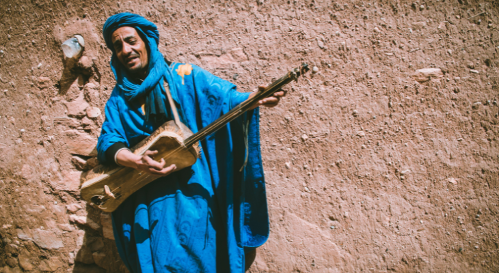 Man in blauwe traditionele kleding speelt snaarinstrument tegen een zandkleurige muur.