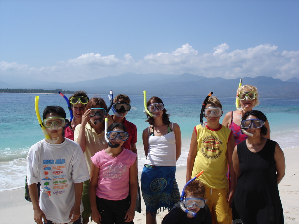 Gezin met snorkelmaskers op het strand tijdens familiereis Java en Bali, met helderblauwe zee en bergen op de achtergrond.