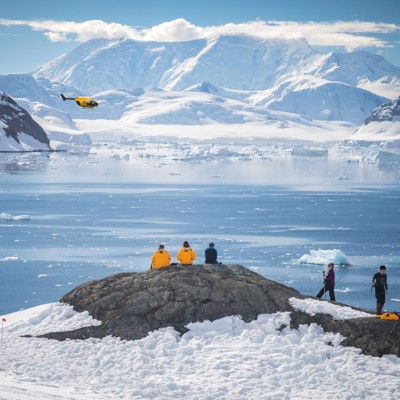 Helikopter-landing bij gletsjer: helikopter vliegt boven ijsfjord terwijl hikers uitkijken over besneeuwd landschap.