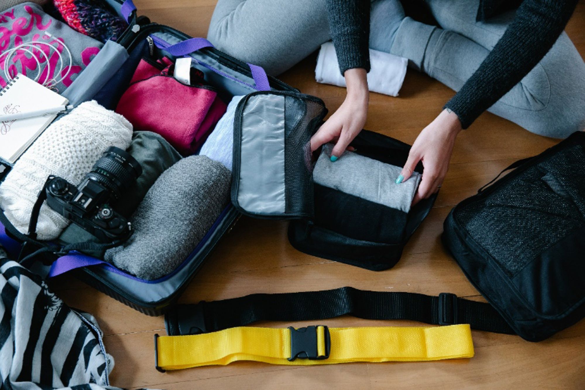 Persoon pakt kleding in een open koffer met camera en reisaccessoires op de vloer.