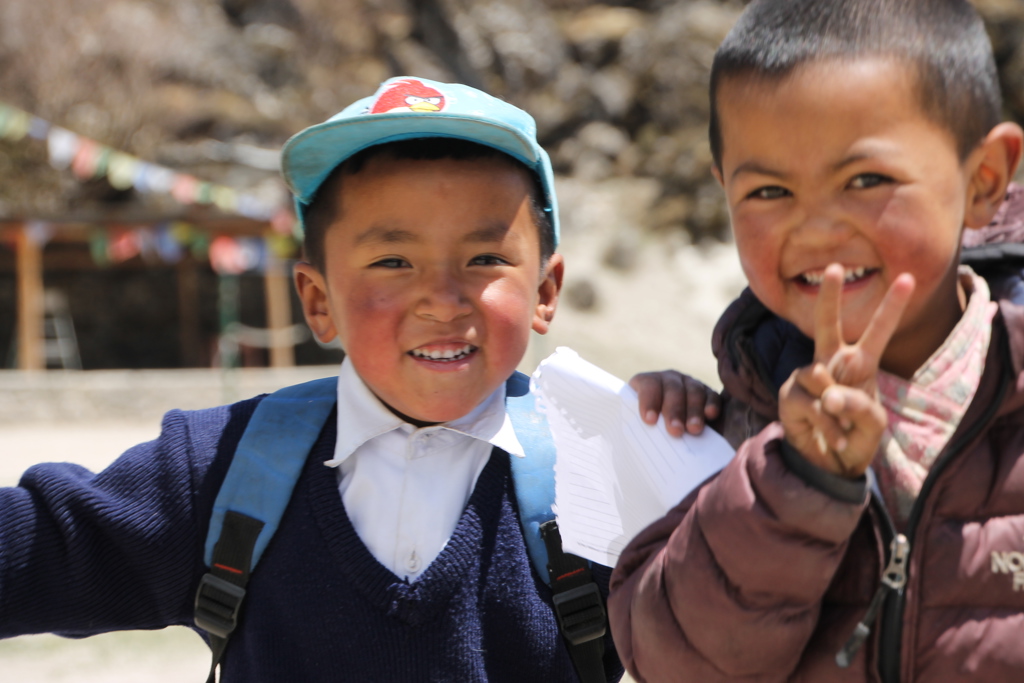 Twee lachende kinderen in Nepal, één met schooltas en pet, tijdens een familiereis.