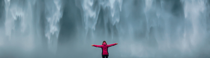 Reiziger in rode jas met gespreide armen voor een enorme waterval, reisaanbevelingen .