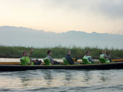 Reizigers in een boot op Inle Lake in Myanmar, met rietvelden en bergen op de achtergrond.