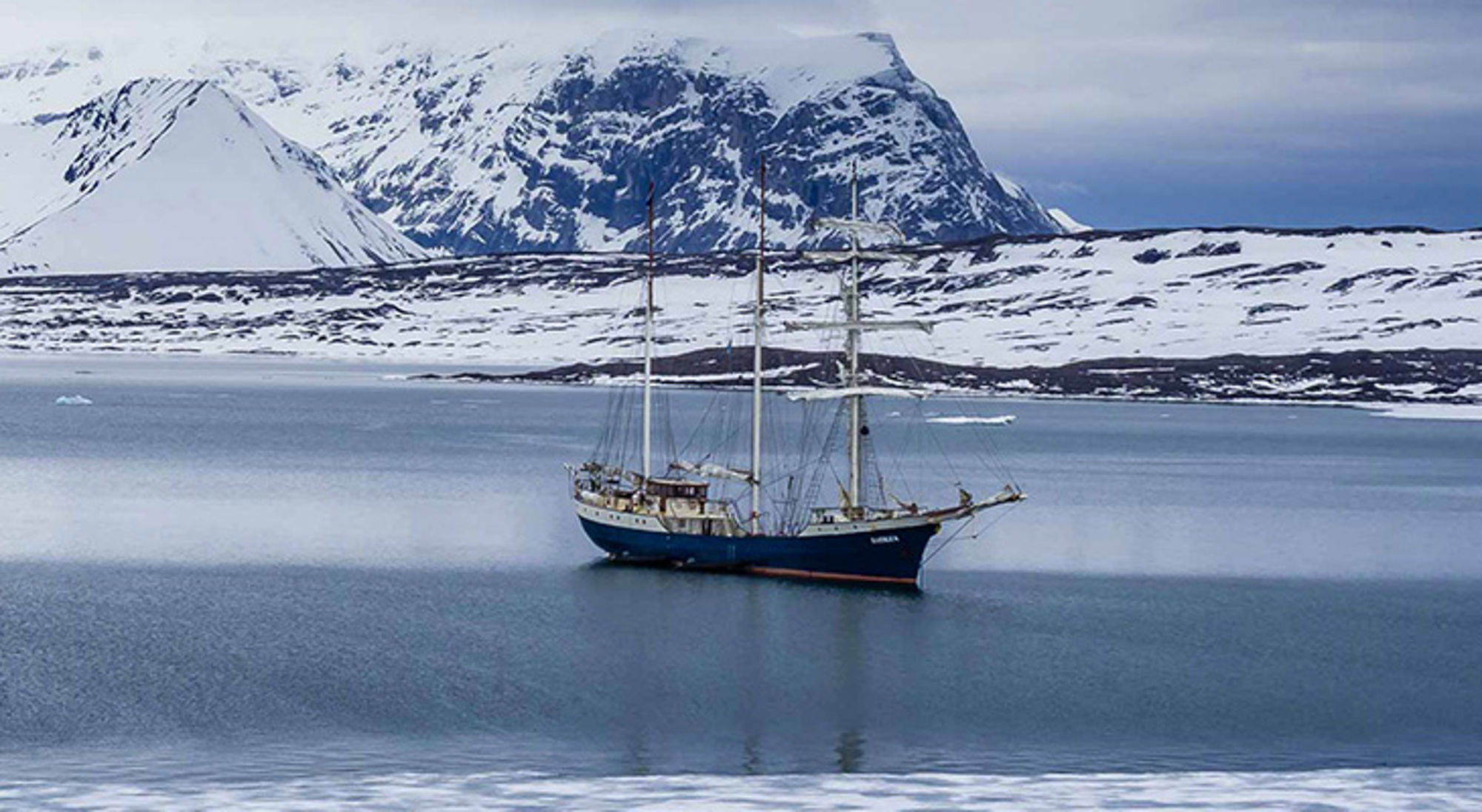 Zeilschip in een rustige fjord met besneeuwde bergen op de achtergrond.