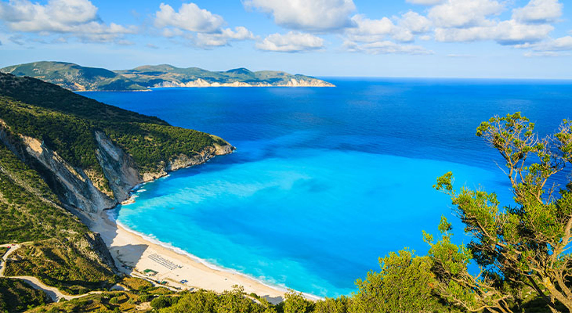 Kefalonia Panoramisch uitzicht op een turquoise baai met zandstrand, steile kliffen en groene heuvels onder een blauwe lucht.