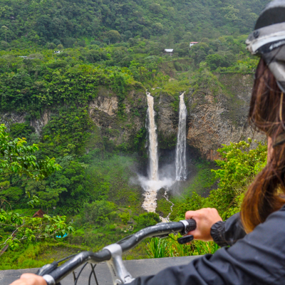 Fietser met helm kijkt uit over een dubbele waterval in het groene berglandschap van Ecuador.