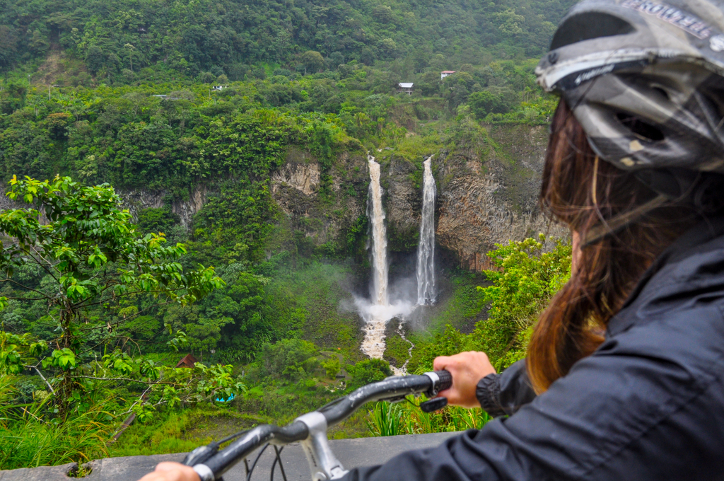 Fietser met helm kijkt uit over een dubbele waterval in het groene berglandschap van Ecuador.