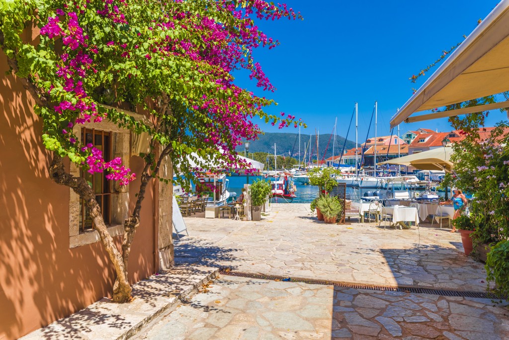 Haven in Griekenland met zeilboten, terrasjes en bougainvillea langs een zonnige boulevard.